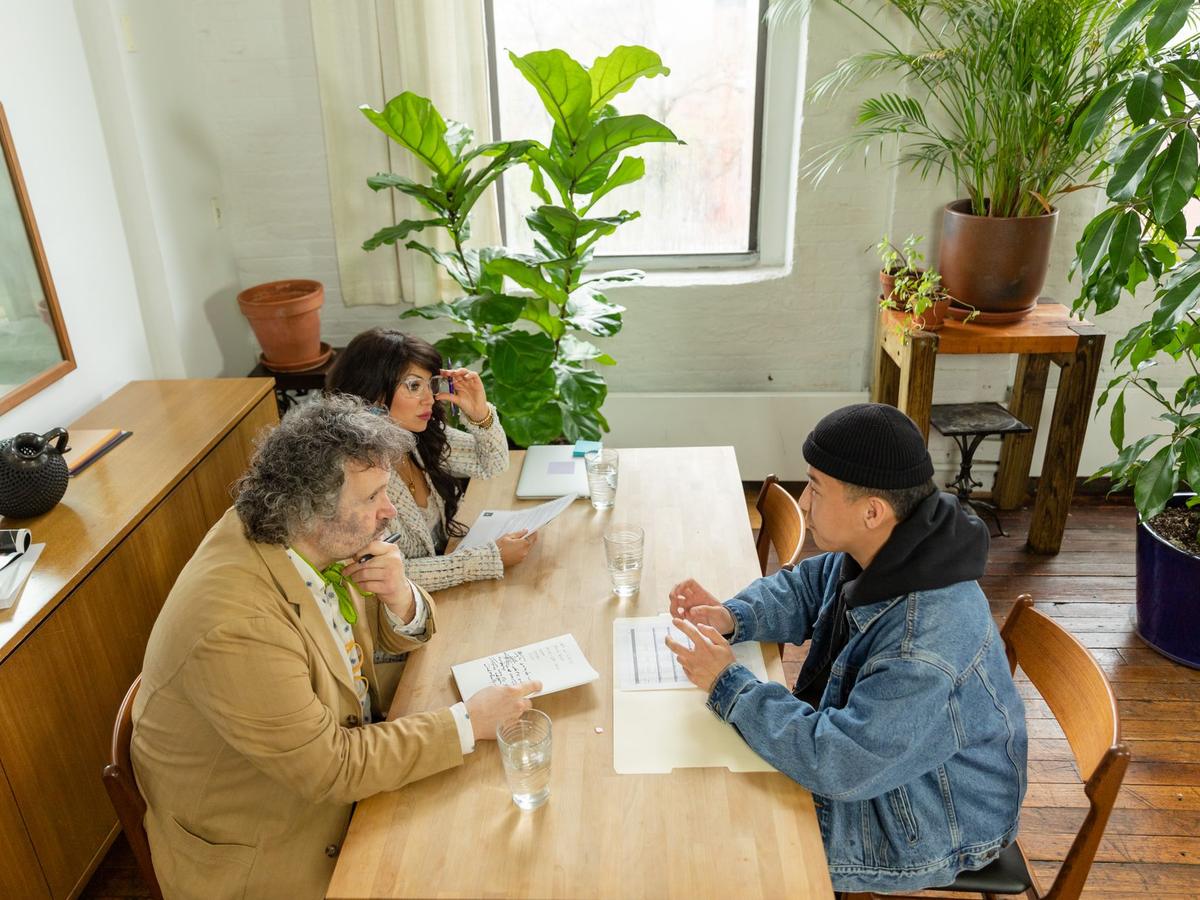 Three colleagues talking over notes at a long wooden table in a plant-filled office